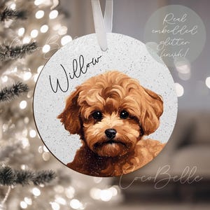 May include: A circular, white Christmas ornament with a glitter effect, displaying a brown dog illustration. The dog's name, "Willow," is written in cursive. A white ribbon is attached. Text reads "Real embedded glitter finish!"
