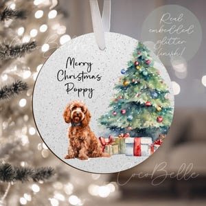 May include: Circular Christmas ornament featuring a brown dog, Christmas tree, and presents. The ornament reads "Merry Christmas Poppy". It has a white ribbon for hanging. The ornament has a glitter finish.