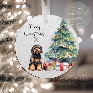 May include: A circular Christmas decoration with a glitter finish. It features a watercolour illustration of a dog, a Christmas tree, and wrapped presents. Text reads "Merry Christmas Ted". A white ribbon is attached for hanging.