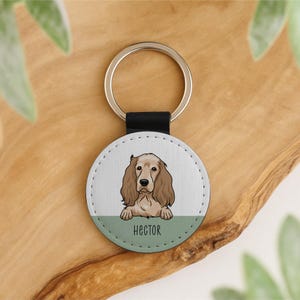 May include: A round keychain with a dog illustration and the name "Hector". The keychain has a silver ring and a black leather strap. The dog is light brown and tan, with a green stripe at the bottom. The keychain is a pet-themed accessory.