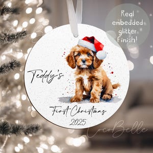 May include: A circular Christmas decoration with a watercolour illustration of a brown puppy in a Santa hat. The ornament reads "Teddy's First Christmas 2025" and has a white ribbon for hanging.