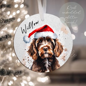 May include: Circular Christmas decoration with a watercolour illustration of a dog wearing a Santa hat. The dog is brown and white with a black nose. The ornament has the name "Willow" written on it. It has a white ribbon for hanging.