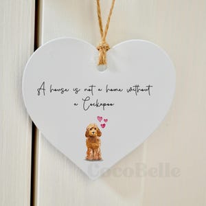 May include: White heart-shaped plaque with a brown string. The plaque has the text "A house is not a home without a Cockapoo" and a watercolour illustration of a Cockapoo dog with hearts.