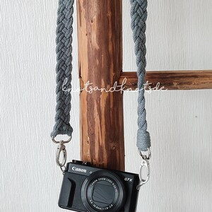 Puede incluir: Correa de cámara trenzada gris con cierres metálicos plateados, sujeta a una cámara Canon G7X negra. La correa cuelga de un poste de madera. El fondo es una pared blanca texturizada.