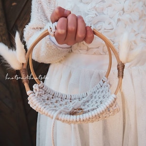 Op de afbeelding: Een handgemaakte macrame ringmand met een houten handvat, perfect voor bruiloften. De mand heeft een geweven ontwerp en is versierd met gedroogde witte bloemen en een kleine ring. De tekst "knotsandknotsde" is zichtbaar.