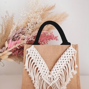 Könnte beinhalten: Eine natürliche Tragetasche aus Jute mit schwarzem Griff und cremefarbenem Makramee-Design. Das Makramee hat eine V-Form mit Fransen. Getrocknete Blumen in Rosa, Beige und Braun sind im Hintergrund.