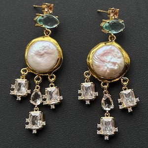 Peut inclure: Une paire de boucles d'oreilles pendantes dorées. Chaque boucle d'oreille présente une grande perle ronde blanc cassé sertie dans un cadre doré. En dessous, des cristaux clairs rectangulaires et en forme de goutte pendent. Les boucles d'oreilles sont ornées de petites pierres précieuses colorées.