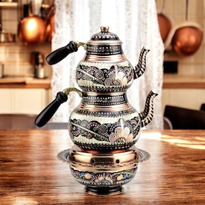 May include: A traditional Turkish tea set with a copper base and two black and white enamel teapots with floral designs. The teapots have black wooden handles.