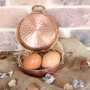 May include: Two brown eggs in a copper pot with a hammered finish. The pot has two brass handles and is sitting on a hessian surface.
