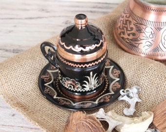 May include: A black and copper Turkish coffee pot with a floral design on the side. The pot is sitting on a burlap surface.