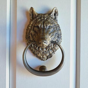 Puede incluir: Un aldaba de puerta con forma de cabeza de lobo en color bronce, con un asa circular. El pelaje y los rasgos faciales del lobo son detallados. La aldaba está montada en una puerta blanca.