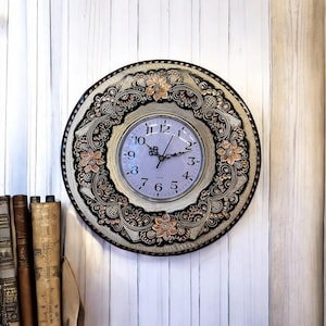 Pode incluir: Um relógio de parede redondo, preto e branco com um design floral de cobre. O mostrador do relógio é branco com números e ponteiros pretos.