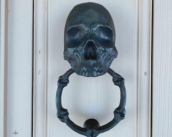 May include: A dark gray skull-shaped door knocker with a ring-shaped handle made of bone-like elements. The knocker is mounted on a white wooden door, creating a striking contrast. The skull features detailed facial features.