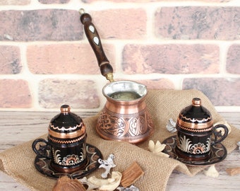 May include: Two black and copper Turkish coffee cups with saucers and a copper coffee pot with a wooden handle. The cups and pot are decorated with intricate floral designs.
