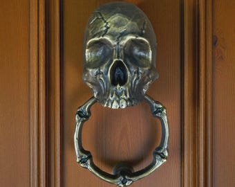 May include: A bronze-colored skull-shaped door knocker with a ring-shaped handle. The skull has detailed features, including eye sockets, a nose cavity, and teeth. The handle is designed to resemble a skeletal hand holding a ring. The knocker is mounted on a wooden door.