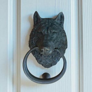 Op de afbeelding: Een gedetailleerde, donker metalen deurklopper in de vorm van een wolfskop. De wolfskop heeft ingewikkelde vachtdetails en houdt een cirkelvormige handgreep in zijn bek. De klopper is gemonteerd op een witte houten deur.