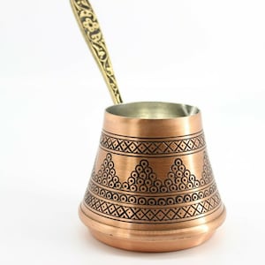 May include: A copper coffee pot with a decorative black design. The pot has a long, ornate handle.