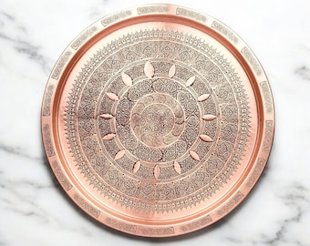 May include: A round copper serving tray with an intricate engraved design. The tray has a raised rim and a hammered finish.