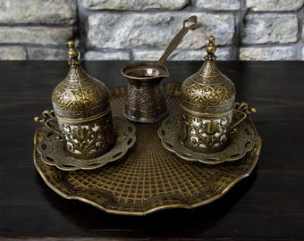 May include: A set of two ornate gold and white Turkish coffee cups and a matching pot on a decorative tray. The cups and pot have intricate floral designs.