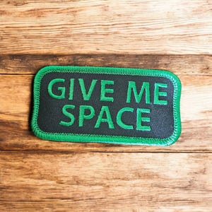 May include: A rectangular patch with the text "GIVE ME SPACE" embroidered in green on a black background. The patch has a green border and is set against a wooden background.
