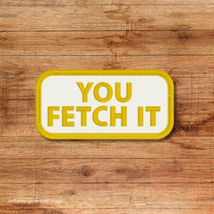 May include: A rectangular patch with the text "YOU FETCH IT" in yellow, outlined in yellow, on a white background. The patch has a yellow border and is set against a wooden background.