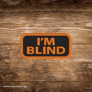 May include: A rectangular patch with the text "I'M BLIND" in orange letters on a black background. The patch has an orange border and is set against a wooden background.