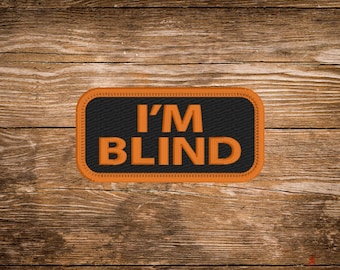 May include: A rectangular patch with the text "I'M BLIND" in orange letters on a black background. The patch has an orange border and is set against a wooden background.
