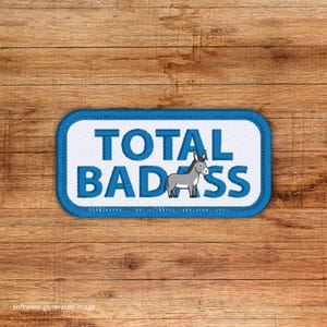 May include: A rectangular patch with a blue border and the words "TOTAL BADASS" in bold white letters. A gray donkey illustration is centered below the text. The patch is on a wooden surface.