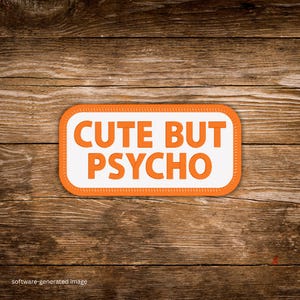 May include: A rectangular patch with the words "CUTE BUT PSYCHO" in orange block letters on a white background. The patch has an orange border and is set against a wooden background.