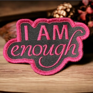 May include: A black patch with pink embroidered text that reads "I AM enough." The patch has a pink border and a cloud-like shape. The patch is on a wooden surface.