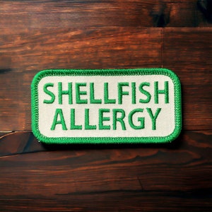 May include: White rectangular patch with a green border. The text "SHELLFISH ALLERGY" is embroidered in green.