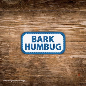 May include: A rectangular patch with the words "BARK HUMBUG" in blue block letters on a white background. The patch has a blue border with white stitching and is set against a wooden background.