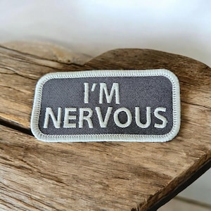 May include: A rectangular patch with the words "I'M NERVOUS" embroidered in white on a dark gray background. The patch has a light gray border and is displayed on a piece of weathered wood.