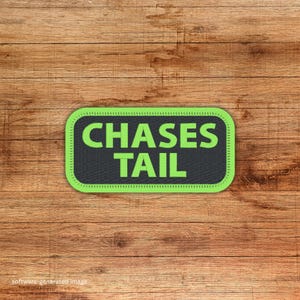 May include: A rectangular patch with the words "CHASES TAIL" in bright green letters on a black background. The patch has a green border with a dotted pattern. The patch is set against a wooden background.