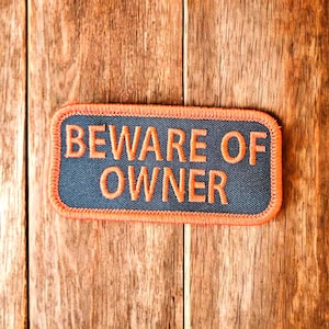 May include: A black and orange embroidered patch with the text "BEWARE OF OWNER" on a wooden background.