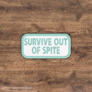 May include: A rectangular patch with the words "SURVIVE OUT OF SPITE" in light blue, outlined in a matching color. The patch has a white background and a light blue border. The patch is set against a wooden background.