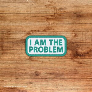 May include: A rectangular patch with the text "I AM THE PROBLEM" in white block letters on a teal background. The patch has a white border and a teal stitched edge. The patch is set against a wooden background.