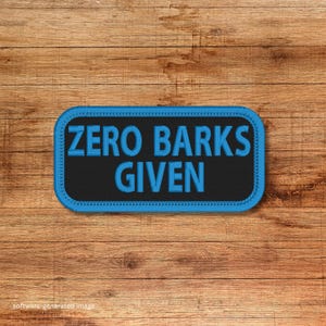 May include: A rectangular patch with the text "ZERO BARKS GIVEN" in blue, embroidered on a black background. The patch has a blue border and is set against a wooden background.