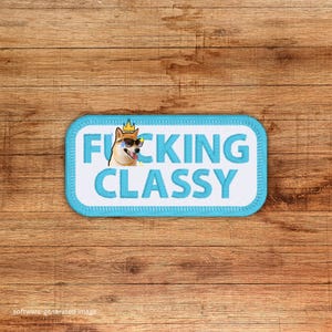 May include: A rectangular patch with a light blue border and the words "FUCKING CLASSY" in bold, light blue letters. A cartoon Shiba Inu dog wearing sunglasses and a crown is in the upper left corner. The patch is on a wooden surface.