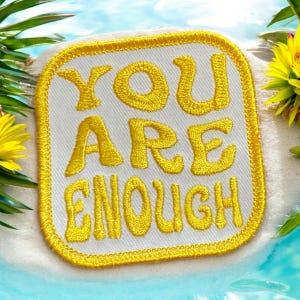 May include: A white square patch with a yellow embroidered border and the text "YOU ARE ENOUGH" in yellow.