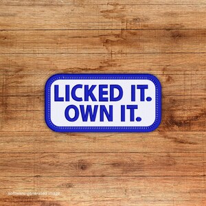 May include: A rectangular patch with the text "LICKED IT. OWN IT." in dark blue, centered on a white background. The patch has a blue border with white stitching. The background is a wooden surface.