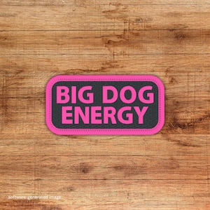 May include: A rectangular patch with the words "BIG DOG ENERGY" in bright pink letters on a black background. The patch has a pink stitched border and is set against a wooden background.