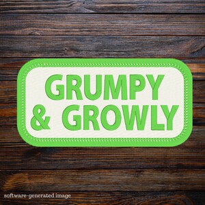 May include: A rectangular patch with rounded corners, featuring the words "GRUMPY & GROWLY" in green, embroidered on an off-white background. The patch is outlined with a green border and set against a dark wood grain backdrop.
