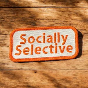 May include: A rectangular patch with the words "Socially Selective" in orange letters. The patch has a white background with an orange border and is set against a wooden surface.
