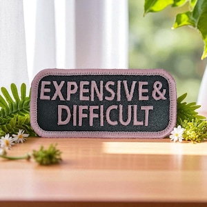 May include: A rectangular patch with the words "EXPENSIVE & DIFFICULT" embroidered in pink on a black background. The patch has a pink border and is set against a wooden surface with greenery.