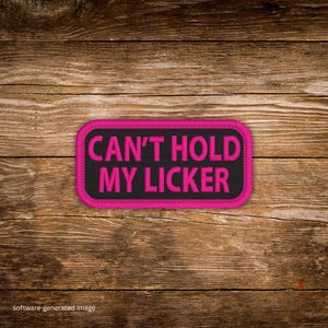May include: A rectangular patch with the text "CAN'T HOLD MY LICKER" in bright pink, outlined in pink stitching, on a black background. The patch is set against a wooden background.