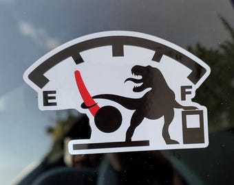 May include: A white sticker with a black outline of a fuel gauge. The gauge shows the fuel level is low, with a red needle pointing towards "E". A black Tyrannosaurus Rex is featured in the center of the gauge.