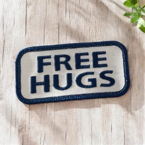 May include: A white fabric patch with a blue border and the text "FREE HUGS" embroidered in blue.