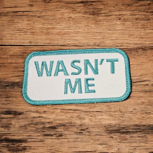 May include: A white patch with a teal border and the text "Wasn't Me" embroidered in teal.