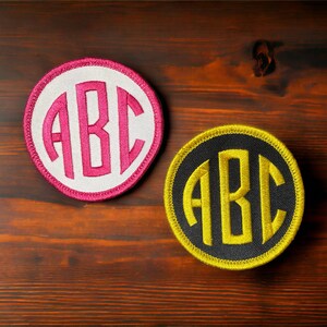 May include: Two round fabric patches with embroidered white and black circles containing the letters "ABC" in pink and yellow thread.
